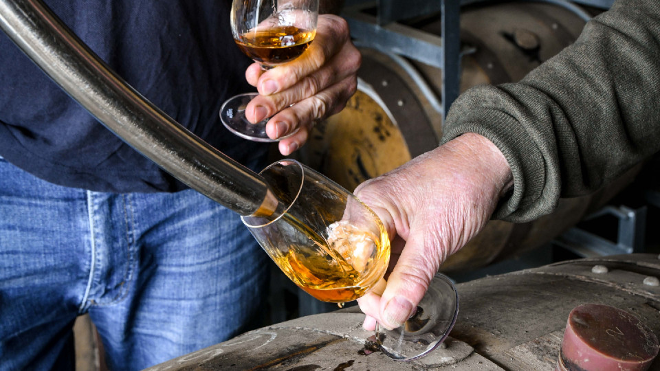 A hand holding a glass over a barrel as spirits are poured into it.