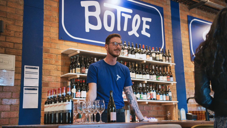 A bartender stands at the Bottle Shock bar. He's wearing a blue company t-shirt. Various wine bottles sit on the bar in front of him and on the shelves behind.